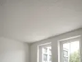 Jasny, nierówny sufit pokryty farbą strukturalną. Widać dwa okna z widokiem na budynki i drzewa.