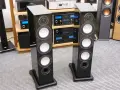 Dwie eleganckie kolumny głośnikowe Monitor Audio RX8 w czarnym połysku, stojące na szarym dywanie, obok sprzętu audio.
