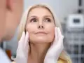 Kobieta z blond włosami poddaje się zabiegowi depilacji laserowej twarzy. Dłonie w rękawiczkach delikatnie dotykają jej policzków.