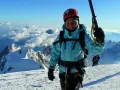 Barbara Prymakowska, mimo swojego wieku, zdobywa szczyty. Uśmiechnięta alpinistka w kasku i turkusowej kurtce na tle gór i chmur.