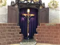 Kreuzweg Stationen: Jesus am Kreuz, umgeben von Ziegelmauern, die die "Mauer der Klage" symbolisieren. Eine Kerze brennt vor dem Kruzifix.