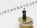 Miniatur-Priester steht auf Münzstapel. Der Schriftzug "Kirchensteuer" im Hintergrund wirft die Frage auf, warum man kirchensteuer zahlt.