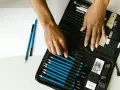 Un étui ouvert révèle une sélection de crayons de différents types pour dessiner, avec des mains choisissant un crayon bleu.