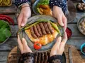 Ręce trzymają talerz z grillowanymi **ćevapi** na płaskim chlebie, ozdobiony papryczkami i pomidorem. Obok sałatka i inne dania.