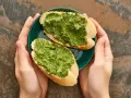 Dwie kromki bagietki z aromatycznym pesto z pietruszki na zielonym talerzyku, trzymanym w dłoniach.