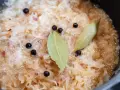 Sauerkraut köchelt mit Lorbeerblättern und Wacholderbeeren, um den Geschmack zu verfeinern. Kleine rote Zwiebelstücke sind ebenfalls sichtbar.