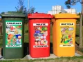 Trzy kosze na śmieci: zielony (kompost), czerwony (odpady) i żółty (recykling), z naklejkami pokazującymi przykłady wyrzucanych rzeczy.