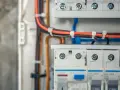 Szczegółowy widok instalacji elektrycznej w domu: przewody w kolorach czarnym i pomarańczowym, zabezpieczone opaskami, prowadzą do białej skrzynki z bezpiecznikami.