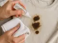 Manos aplicando líquido a una mancha de grasa en ropa beige, usando un paño blanco para frotar. Aprende como quitar grasa de la ropa.