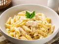 Pyszny makaron rurki w kremowym sosie z kurczakiem, posypany parmezanem i udekorowany natką pietruszki.