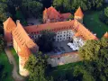 Zamek w Bytowie otoczony zielenią, z czerwonymi dachami i okrągłymi wieżami. Na dziedzińcu widać stoliki i parasole.