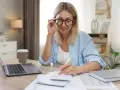 Femme aux lunettes calculant, laptop et café à proximité. Elle semble gérer des finances, peut-être une réduction de capital non motivée par des pertes.