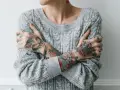 Osoba w szarym swetrze obejmuje się ramionami, ukazując kolorowe tatuaże na przedramionach i dłoniach. Widać tatuaż kota, róże i czaszkę. To może być przykład, jak wygląda zakażenie tatuażu, gdy skóra jest podrażniona.