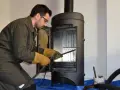 Un homme en tenue de travail effectue le ramonage d'un poêle à bois, ganté et muni d'outils.