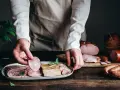 Ręce układają na talerzu plastry pieczeni i wędlin, obok leżą inne włoskie wędliny, tworząc apetyczną kompozycję.