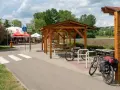 Miejsce odpoczynku przy ścieżce rowerowej. Dwa rowery, w tym jeden z sakwami, czekają na dalszą podróż. W tle zadaszenie i stoliki, idealne na przerwę w trasie.