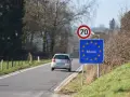 Znak graniczny Belgii, gdzie potrzebny paszport, na tle drogi i jadącego samochodu.