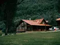 Drewniany domek w górach, idealne miejsce na letni weekend. Dwa samochody zaparkowane przed domem, otoczonym zielenią i lasem.