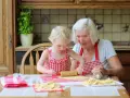 Babcia z wnuczką pieką ciasteczka. Dziewczynka wałkuje ciasto, a babcia z uśmiechem patrzy na jej pracę.