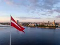 Flaga Łotwy powiewa nad rzeką w Rydze, ukazując panoramę miasta o zachodzie słońca.