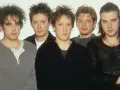Pięciu członków zespołu The Cure pozuje do zdjęcia. Robert Smith z charakterystyczną fryzurą i makijażem na pierwszym planie.