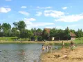 Sonniger Tag am badesee fredenbeck. Kinder planschen im Wasser und am Sandstrand, während im Hintergrund Gebäude und Bäume zu sehen sind.