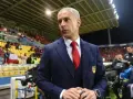 Sylvinho, trener w granatowym garniturze i czerwonym krawacie, stoi na tle trybun stadionu z flagą Chile.