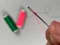 Dedos sujetan una aguja para meter hilos, con carretes de hilo verde y rosa desenfocados al fondo.