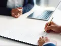 Persona firma un contrato de alquiler de habitación. A su lado, otra persona espera, con un portátil y una tablet sobre la mesa.