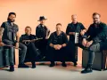 Sechs Männer einer Band posieren vor orangefarbenem Hintergrund, einige sitzen, einer steht.