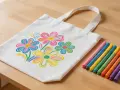 Un sac en tissu blanc décoré de fleurs colorées, réalisé avec de la peinture acrylique. Des feutres de différentes couleurs sont disposés à côté.