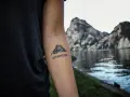 Tatuaż góry na przedramieniu z napisem "ADVENTURE". Symbolizuje miłość do podróży i odkrywania nowych miejsc.