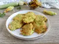 Buñuelos de calabacín dorados y crujientes en un plato blanco, con calabacines y flores en el fondo.