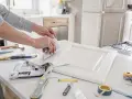 Ręce wygładzają wzorzystą okleinę na białej, frezowanej ramie meblowej. Narzędzia i taśma na blacie.