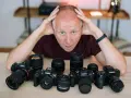 Mężczyzna z rękami na głowie otoczony aparatami różnych marek: Sony, Canon, Fujifilm.