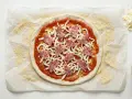 Gotowe ciasto na pizzę bez wyrastania, posmarowane sosem, posypane serem i salami. Obok miseczka sosu i dodatkowy ser.