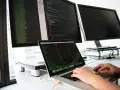 Hands typing on a laptop, surrounded by monitors displaying code. This setup is ideal for developing network infrastructure software.