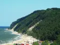Słoneczna plaża u podnóża zalesionej Kawczej Góry. Woda faluje, a na piasku odpoczywają ludzie.
