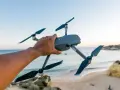 A hand holds a drone over a European beach, ready for flight.