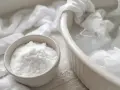 Un bol de bicarbonate de soude à côté d'un plat rempli d'eau et de linge blanc. Idéal pour éliminer les taches jaunes.