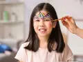 Ein Kind wird mit einem bunten Regenbogen-Einhorn-Gesicht bemalt. Ein Pinsel wird sanft über die Stirn geführt.