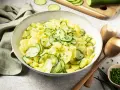 Schüssel mit Kartoffelsalat mit Gurken, garniert mit Schnittlauch. Daneben liegen Servierbesteck und eine kleine Schale mit gehacktem Schnittlauch.