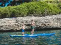 Zwei Jungen auf SUP-Boards, die geradeaus fahren, genießen das klare Wasser vor einer felsigen Küste mit üppiger Vegetation.
