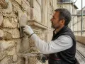 Un artisan applique du mortier sur un mur en pierre pour rénover une maison ancienne. L'échafaudage indique que le travail est en cours.