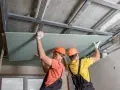 Dwóch budowlańców w kaskach montuje płyty gipsowo-kartonowe do konstrukcji podwieszanego sufitu.