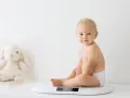 Uśmiechnięty chłopczyk w pieluszce siedzi na wadze. Chyba już czas na większy rozmiar, kiedy pampers jest za mały.