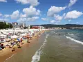 Tłum ludzi wypoczywa na piaszczystej plaży Słoneczny Brzeg, opalając się na leżakach pod białymi parasolami. W tle widać morze i zabudowania kurortu.