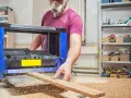 Stolarz w masce ochronnej pracuje przy strugarce, wygładzając drewno. Wióry sypią się wokół.