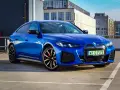 Niebieskie BMW i4 M50 zaparkowane na parkingu, z widokiem na budynki w tle.