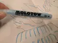 A hand holds a light blue Molotow Grafx Art Masking Liquid Pump Marker over a drawing.